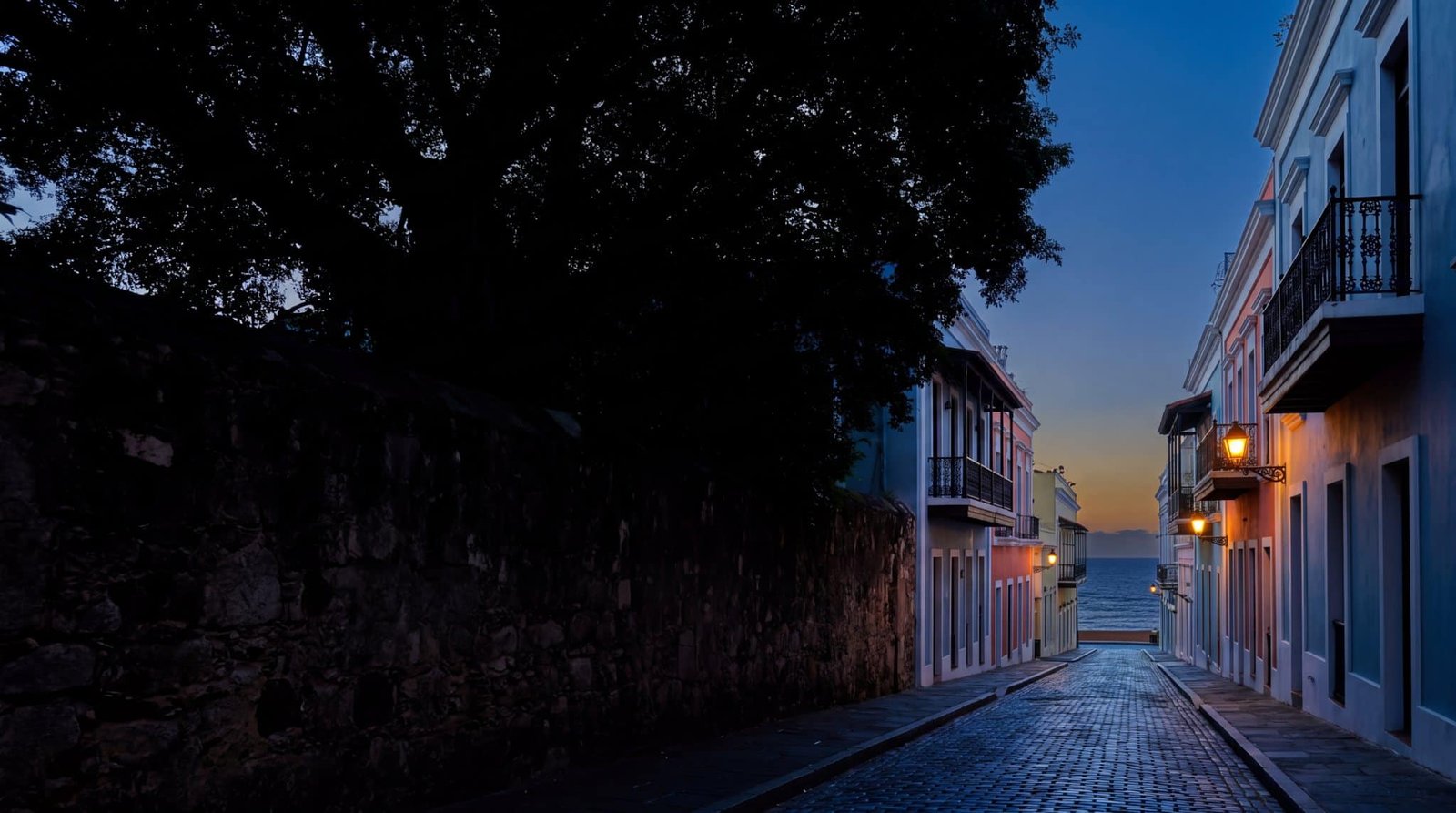 A lamplit cobblestone street of pastel colonial houses in Old San Juan running downhill to the Atlantic at dusk